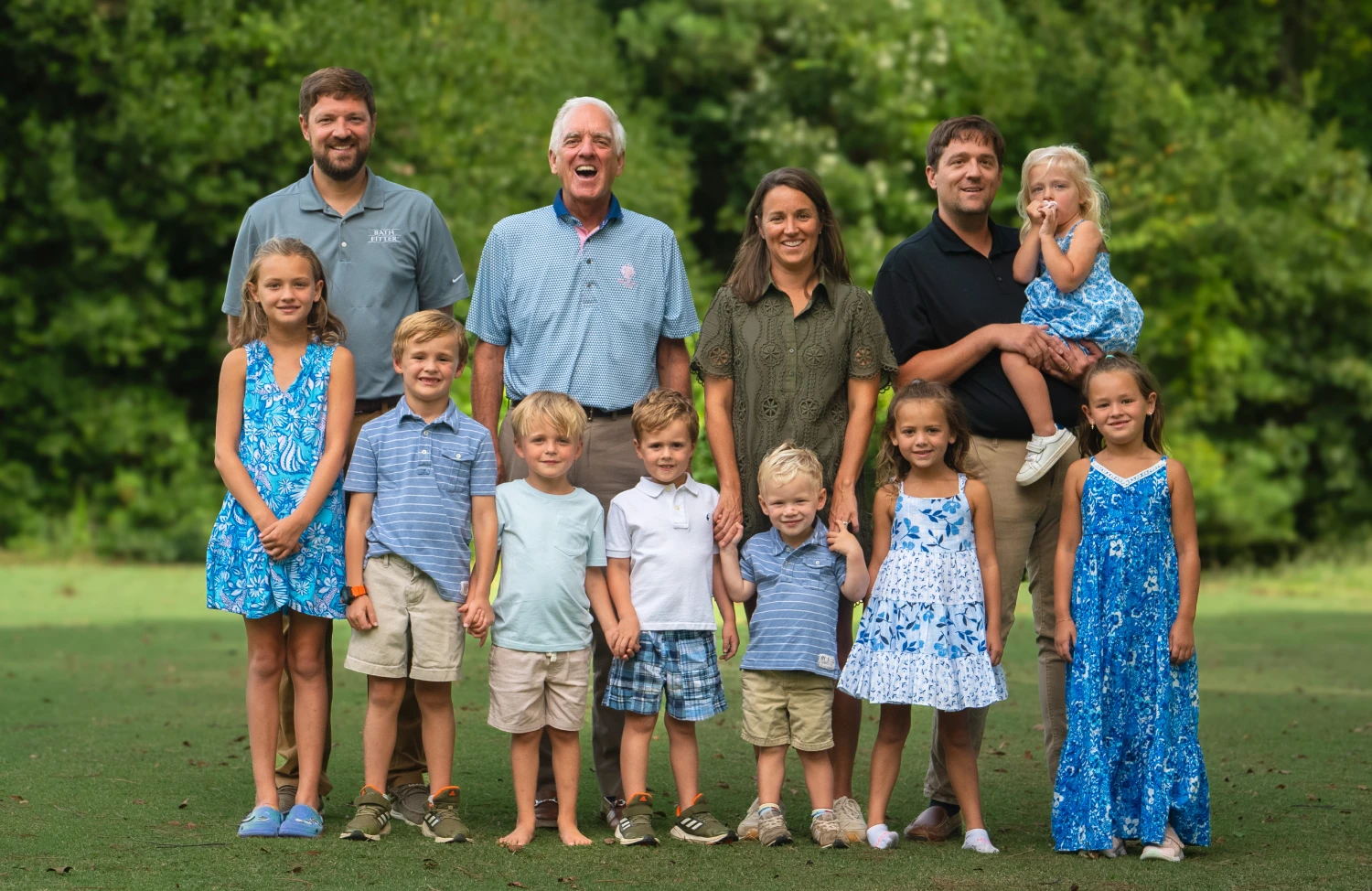 A large family portrait of three generations standing on a grassy lawn with a lush green background. The group includes three adult men, two adult women, and nine children of various ages. The adults are smiling, and the children are positioned in front and around them.